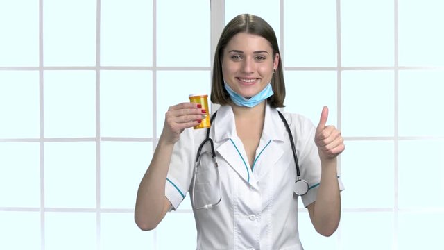 Portrait of doctor with approval sign. Happy female doctor holding container with pills and gesturing thumb up. Concept of success.