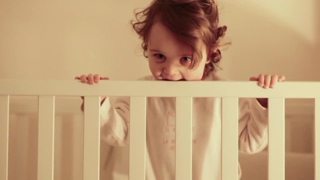 Toddler baby girl bites edge of the cot.
