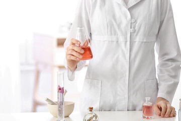 Woman in lab coat holding glass flask with perfume oil near table indoors