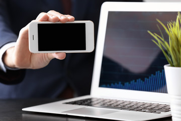 Man holding smartphone near laptop with stock chart data on screen. Forex concept