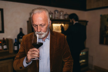 Mature sommelier smelling flavor of red wine in glass, he is checking color, quality and sediments of drink. 