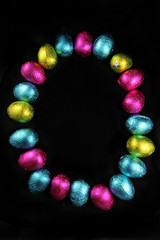 Colorful foil wrapped easter eggs laid out in the oval shape of an egg, on a black background.