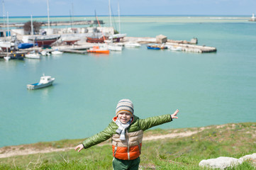 dreaming little child in jacket and hat stands on green hill spreading his hands like wings on background of sea bay with boats in cold spring