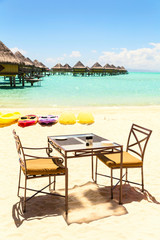 Dining table with two chairs on sandy beach by tropical sea side, with colourful kayaks and water villas background