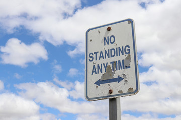 a weathered, peeling blue and white No Standing Any Time sign with double ended arrow in a cloudy sky