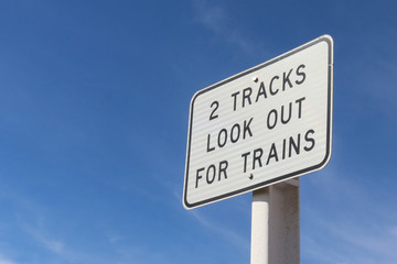 a black and white 2 Tracks Look Out For Trains warning sign in a blue sky