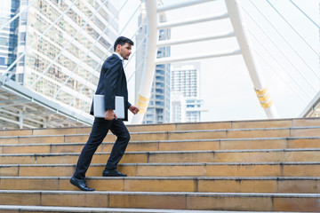 Business man celebrating success. Young businessman walking at stairs and hold laptop computer while standing outdoors with office building in the background.