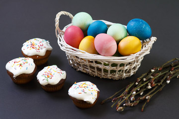 Easter concept. Multicolored Easter eggs in basket, sweet muffins and willow branches on black background. Easter background. Treats for the spring religious Christian holiday