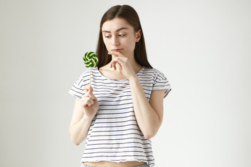 Isolated portrait of thoughtful young brunette woman in striped top holding round hard candy and touching chin with pensive expression, thinking, being uncertain about having unhealthy lollipop