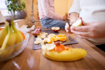Close up of preparation fruit salad and little toddler girl sitting on the kitchen table and waiting while her pregnant mum cutting an apple.