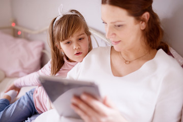 Adorable cute curious girl toddler leaning a head on pregnant mother shoulder and looking at a tablet.