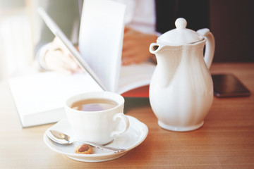 Female hands hold a pen, a book, A white kettle and a cup with a saucer and a teaspoon stand on a table