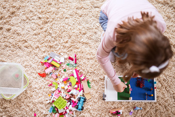 Top view of small toddler girl sitting on the carpet of her room and playing with plastic toys with mess next to her.