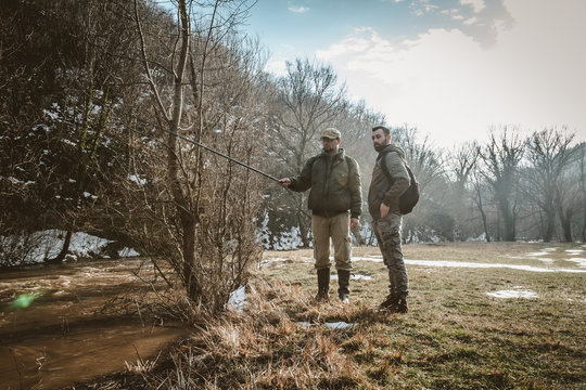 Two young bearded men  standing next to a swollen river, talking and fishing.