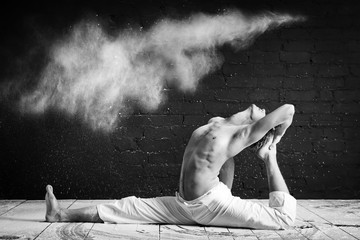 A man doing yoga in a white cloud of dust in a dark room. The concept of energy. Black-and-white photo. Eka pada rajakapo of hanumanasana. pigeon pose