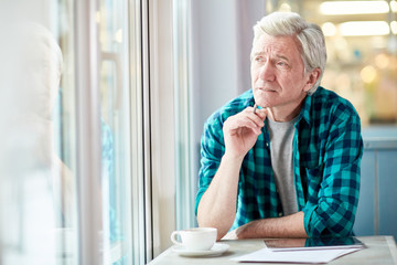 Pensive mature man with tablet and cup of coffee looking through window while getting inspired in cafe