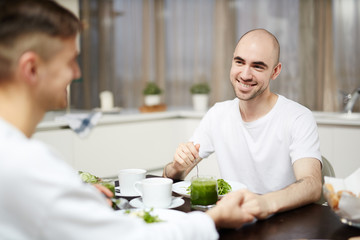 Young cheerful man holding his gay partner by hand while having breakfast in the morning