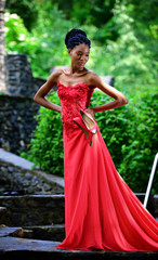 African American girl in a red dress, with dreadlocks, with red shoes in hand, posing in the summer in the Park on a background of green plants. Portrait. Vertical view