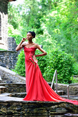 Fototapeta premium African American girl in red dress and with red shoes in her hand, standing with her eyes closed in the summer on the rocks in the Park against the green plants