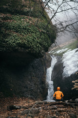 Man sits at the bottom of waterfall in the spring forest 