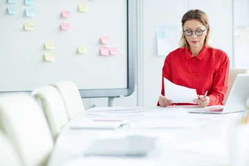 Confident economist in smart casual sitting by table in boardroom and reading business documents
