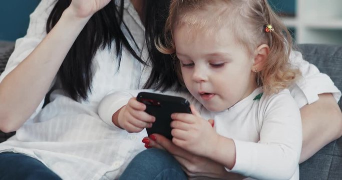 Mom and daughter sit on sofa use phone hug woman baby hand love child person girl kid internet cartoon technology smartphone face touchscreen home mobile looking application touch mother game play
