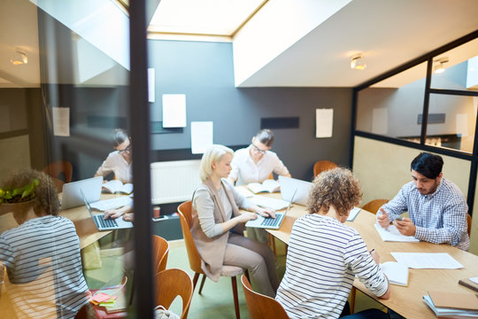 Contemporary young specialists carrying out their working tasks while sitting by long table in office