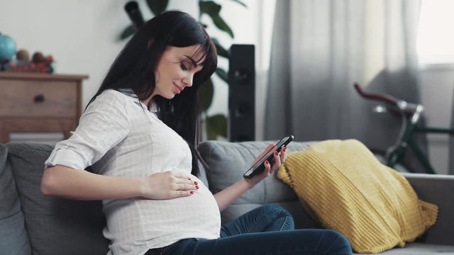 Portrait beautiful pregnant woman use phone sit on sofa touches the abdomen smiling happy smartphone online family home girl internet face technology chatting communication close up attractive