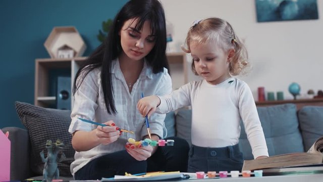Happy mother with little daughter painting paper together smiling kid family hobby baby design love person girl house coloring mom play art paintbrush childhood home table artist closeup talk