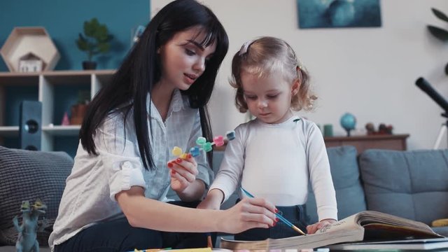 Mom with little daughter painting on paper together hold paintbrushsmiling kid family hobby baby design love person girl house coloring play art childhood happy home table artist closeup talk