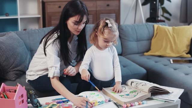 Little child with mother painting on paper book sofa together smiling kid family hobby baby love person girl house coloring mom play art paintbrush childhood happy home table artist closeup talk