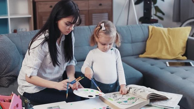 mother with little daughter painting on paper book together smiling kid family hobby baby design love person girl house coloring mom play art paintbrush childhood happy home table artist closeup talk