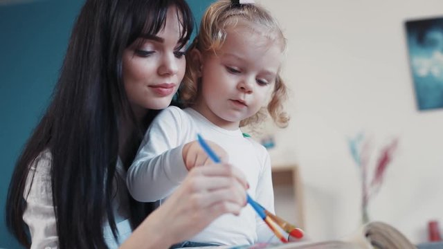 Little child painting on paper book with mum hug sit on sofa together at home happy kid family hobby girl love house coloring mom play art childhood portrait home table artist closeup mother