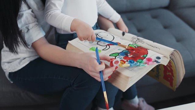 Close up hand little child painting on paper book with mum hug sit on sofa together at home happy kid family hobby girl love house coloring mom play art childhood portrait table artist closeup mother