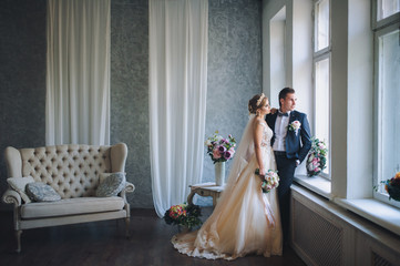 The newlyweds stand at the window in a stylish studio. Beautiful bride in a lace dress.