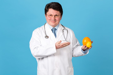 Male doctor holds fruit in hands. Vitamins for your health. Middle aged man doctor in white uniform stretching out lemon and orange fruit, smiling, while standing against blue background.