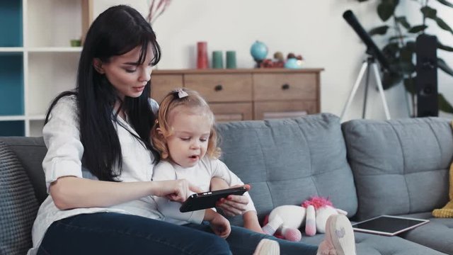 Happy mom and daughter sit on sofa use phone hug woman baby hand love child person girl kid internet cartoon technology smartphone face home mobile looking application touch mother game little play