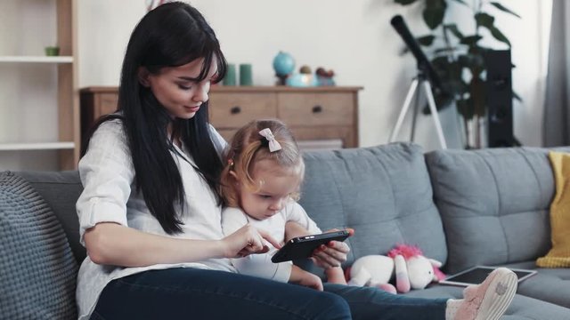 Kid with mother sit on sofa use phone look some interesting hug woman baby hand love child girl internet cartoon technology smartphone face home mobile touch mom game little play application