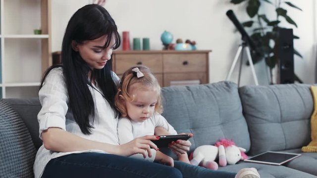 Portrait kid with mother sit on sofa use phone look some interesting hug woman baby hand love child girl internet cartoon technology smartphone face home mobile touch mom game little play application