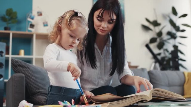 little child hold paintbrush painting on paper book with mum hug sit on sofa together at home happy kid family hobby girl love house coloring mom play art childhood table artist closeup mother