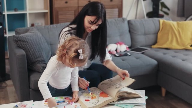 Brunette mother with daugther painting on paper book hug sit on sofa together at home happy kid family hobby girl love house coloring mom play art childhood table artist closeup cute
