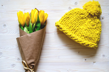 A yellow knitted cap of thick Merino yarn. Nearby is a bouquet of yellow tulips wrapped in paper.
