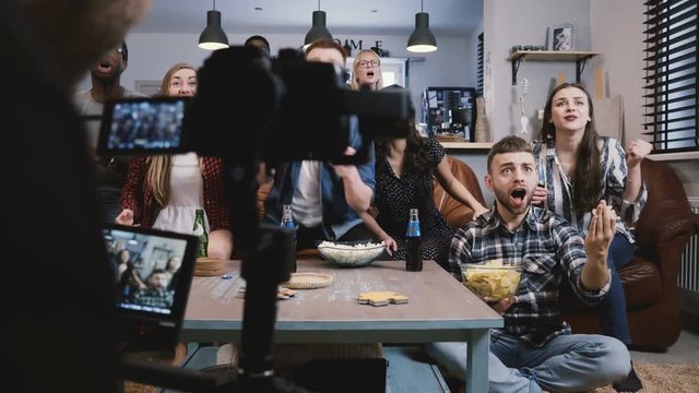 Diverse group of friends watch sports on TV. Backstage camera films happy supporters, fans on couch cheer with snacks 4K