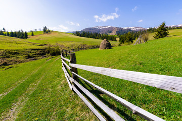 Fototapeta na wymiar haystack behind the wooden fence on a grassy hill. beautiful Carpathian countryside in springtime. mountain ridge with snowy tops in the distance