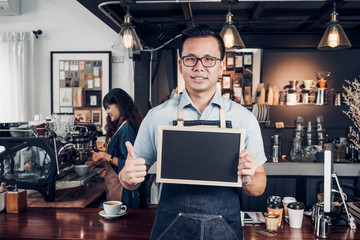 Fototapeta na wymiar Asian male barista wear jean apron thumbs up at blank blackboard coffee menu at counter bar with smile face,cafe service concept,business owner start upLeave space for adding your text