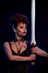 Front view of a beatiful warrior woman keeping steel sword with black skulls. Female wearing bright make up with red lipstick and hairdress with little curles. Confident girl with thin chain on neck.
