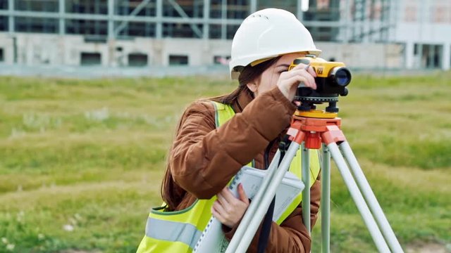 A young woman surveyor in work clothes and helmet adjusts the equipment and produces calculations on the construction site. The concept of landscape design, geodesy