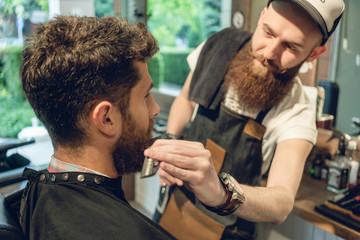Close-up side view of a handsome redhead young man sitting on the chair of a trendy barbershop for trimming and styling his beard