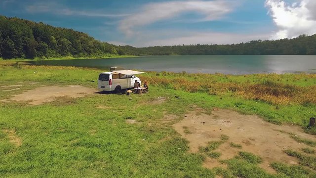 Aerial view circle around white camper van near lake, two people sitting at table under canopy