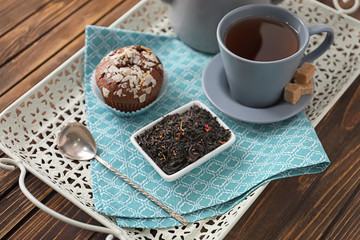 Tray with dry black tea leaves, cup of aromatic beverage and tasty muffin on table